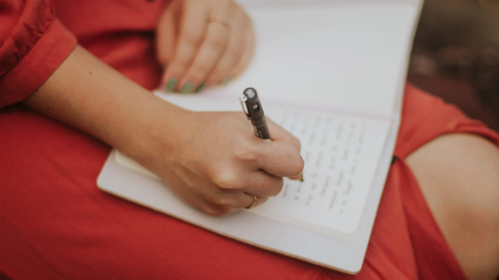 A woman in a red dress writing in an open notebook resting on her lap.