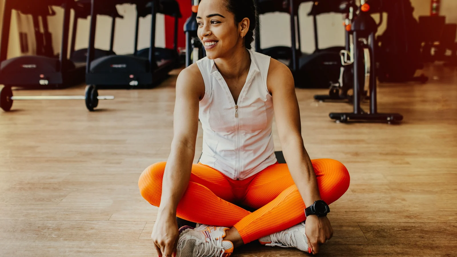 A woman sitting cross-legged on a gym floor in orange leggings, smiling and looking off to one side.
