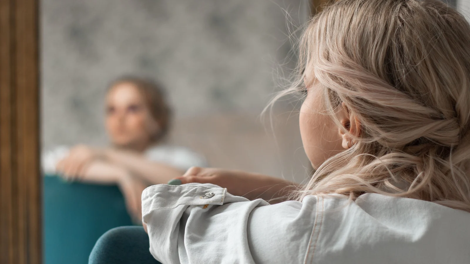 A woman seen from behind, looking at her own reflection in a tall mirror.