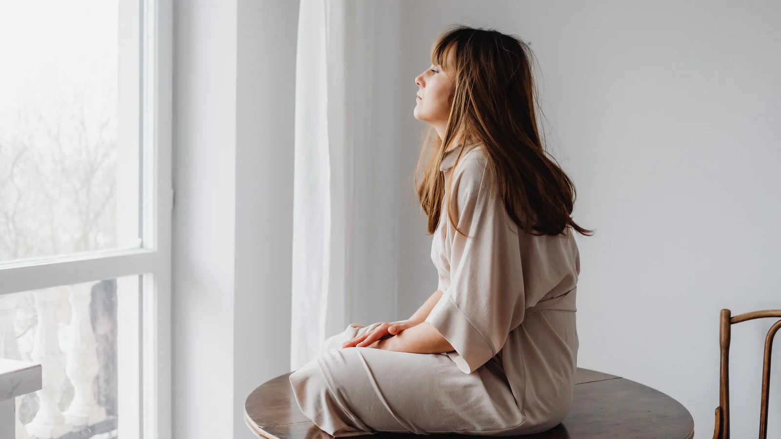 A woman sitting cross-legged on a round table beside a tall window, looking out at the light.