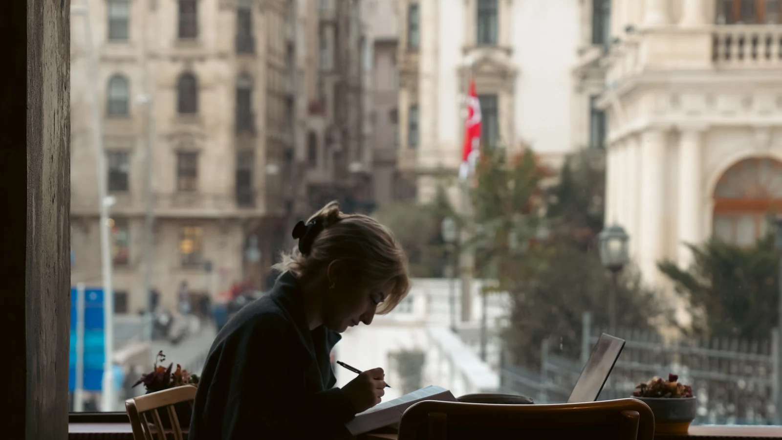 A woman writing in a notebook at a café window, an Istanbul street out of focus behind her.